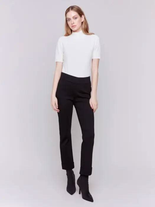 Dressed in stylish Cuffed Ponte Pants by Charlie B and chic black heeled boots, an individual stands with one hand in their pocket, radiating a fashion-forward aura against a plain background.