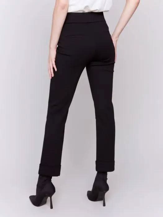 Dressed in stylish Cuffed Ponte Pants by Charlie B and chic black heeled boots, an individual stands with one hand in their pocket, radiating a fashion-forward aura against a plain background.