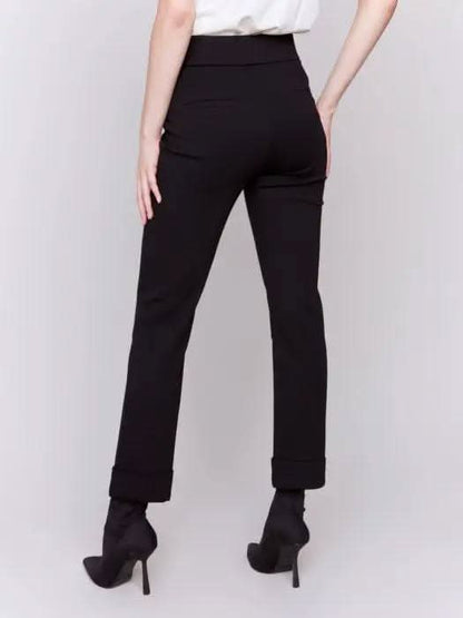 Dressed in stylish Cuffed Ponte Pants by Charlie B and chic black heeled boots, an individual stands with one hand in their pocket, radiating a fashion-forward aura against a plain background.