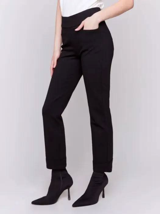 Dressed in stylish Cuffed Ponte Pants by Charlie B and chic black heeled boots, an individual stands with one hand in their pocket, radiating a fashion-forward aura against a plain background.