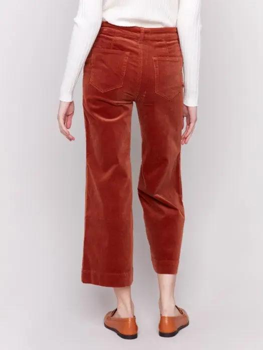 A person wearing a white long-sleeve top, rust-colored Patch Pocket Corduroy Flare Pants from Charlie B made of soft corduroy fabric, and brown loafers stands against a plain background. The high-waisted pants feature a cropped length.