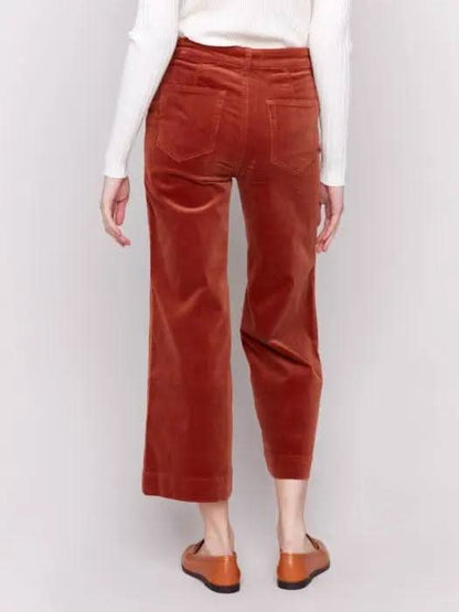 A person wearing a white long-sleeve top, rust-colored Patch Pocket Corduroy Flare Pants from Charlie B made of soft corduroy fabric, and brown loafers stands against a plain background. The high-waisted pants feature a cropped length.