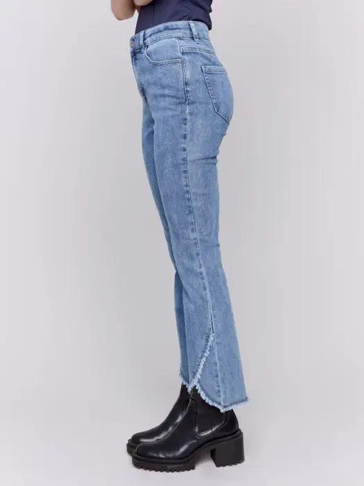 A person wearing Charlie B's light blue Front Tulip Hem Jeans, featuring frayed hem detailing, and black platform boots stands against a light background, showcasing their unique design.