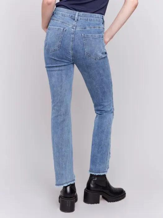 A person wearing Charlie B's light blue Front Tulip Hem Jeans, featuring frayed hem detailing, and black platform boots stands against a light background, showcasing their unique design.
