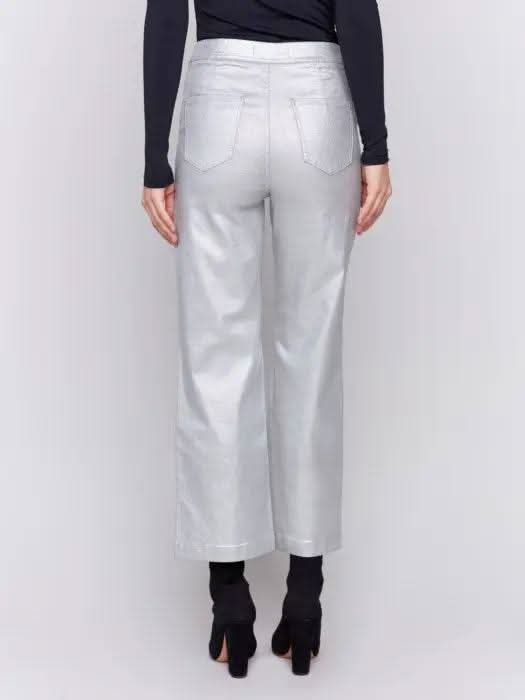 A person sits on a stool wearing a black long-sleeve top, Charlie B's Metallic Wax Flared Pants that shimmer in the light, and black heeled boots against a plain background.