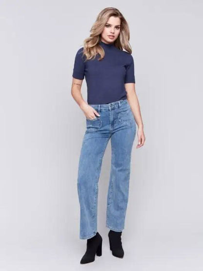 A person wearing a navy blue top, Charlie B flare leg pants with heart-shaped pockets, and black ankle boots stands against a plain gray background.