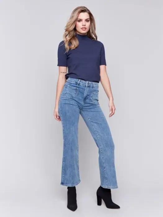 A person wearing a navy blue top, Charlie B flare leg pants with heart-shaped pockets, and black ankle boots stands against a plain gray background.
