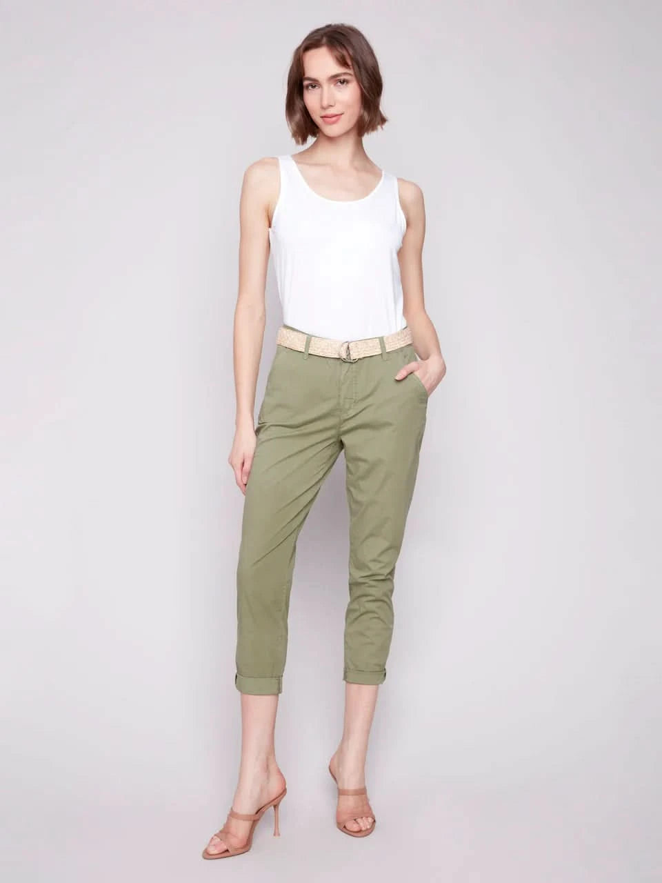 A woman stands against a plain background wearing a white sleeveless top, Charlie B Slim Fit Pants with Belt in celadon green stretch cotton, and nude high-heeled sandals.