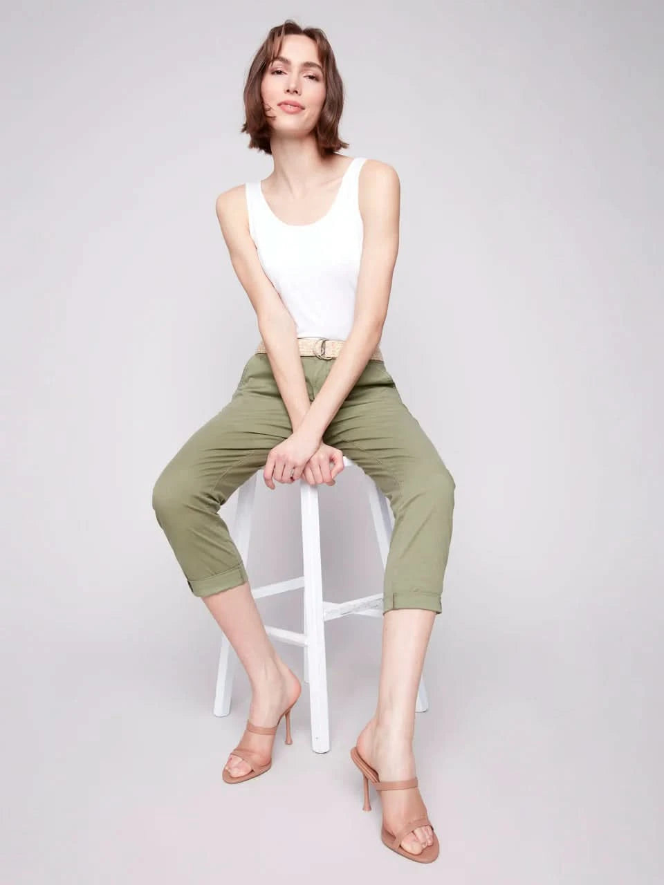 A woman stands against a plain background wearing a white sleeveless top, Charlie B Slim Fit Pants with Belt in celadon green stretch cotton, and nude high-heeled sandals.