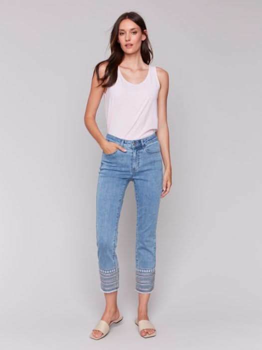 A woman stands against a plain background wearing a sleeveless white top, Charlie B Embroidered Hem Jeans in a slim leg silhouette, and beige sandals, with one hand in her pocket.