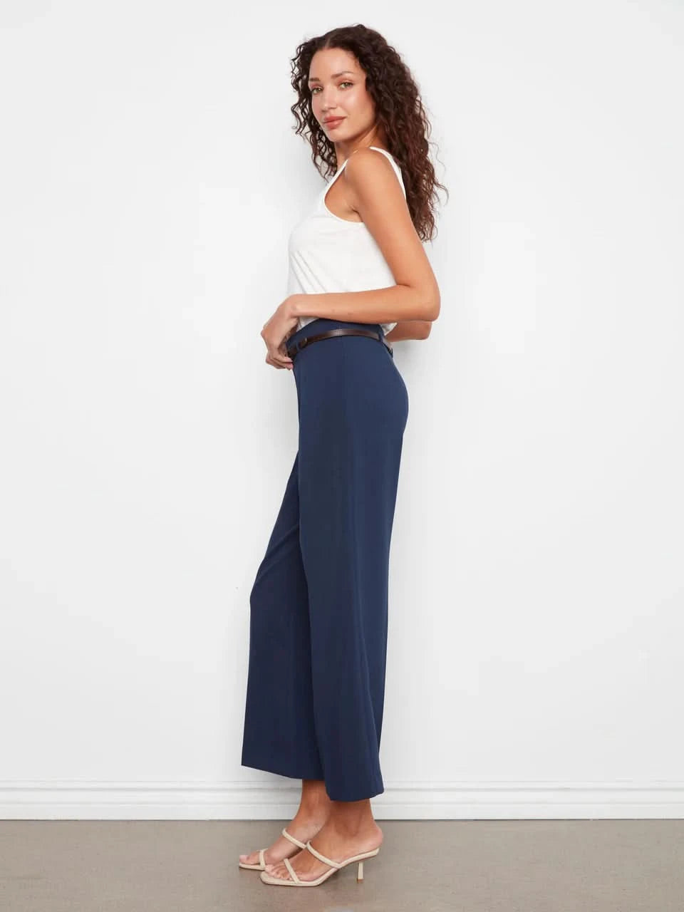 A person stands against a white wall wearing a white sleeveless top, beige heeled sandals, and Charlie B Cropped Dressy Belted Pants in navy blue.