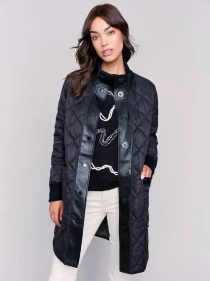 A person with long hair wears a black Charlie B Reversible Quilted Puffer Jacket with a button-up front, a black sweater featuring a white design, and light-colored pants, creating a versatile style perfect for any winter wardrobe. Standing against a plain background, their look is both stylish and adaptable.