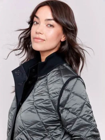 A person with long hair wears a black Charlie B Reversible Quilted Puffer Jacket with a button-up front, a black sweater featuring a white design, and light-colored pants, creating a versatile style perfect for any winter wardrobe. Standing against a plain background, their look is both stylish and adaptable.