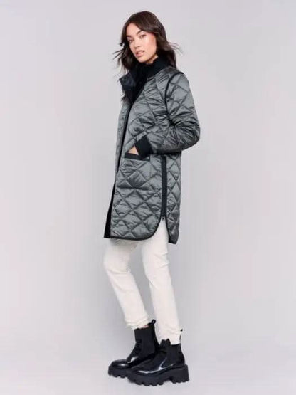 A person with long hair wears a black Charlie B Reversible Quilted Puffer Jacket with a button-up front, a black sweater featuring a white design, and light-colored pants, creating a versatile style perfect for any winter wardrobe. Standing against a plain background, their look is both stylish and adaptable.
