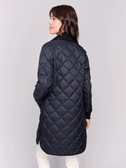 A person with long hair wears a black Charlie B Reversible Quilted Puffer Jacket with a button-up front, a black sweater featuring a white design, and light-colored pants, creating a versatile style perfect for any winter wardrobe. Standing against a plain background, their look is both stylish and adaptable.
