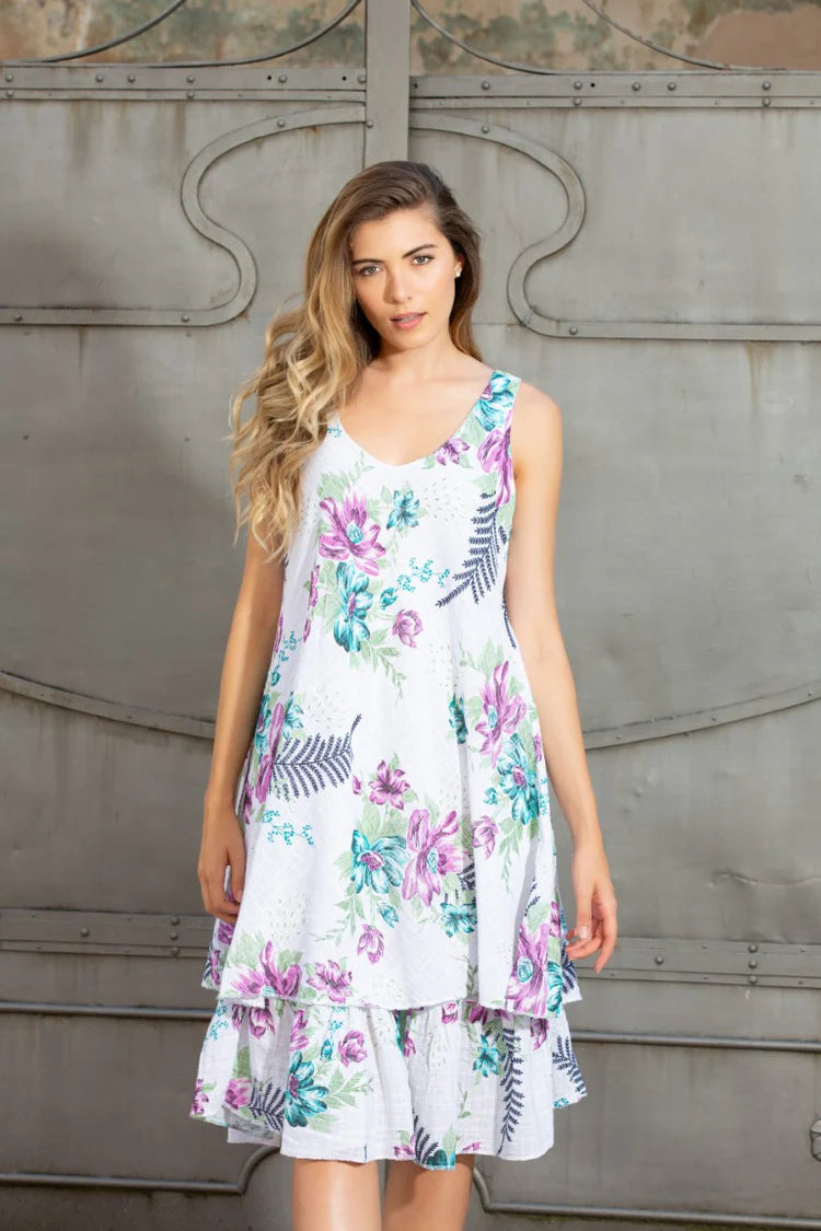 A woman wearing the Variations Isla’s Blooming Breeze Dress, a sleeveless linen dress with a floral print and tiered ruffle hem, stands in front of a decorative metal door.