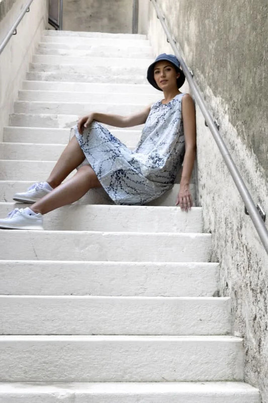 A woman in the Variations Abstract Print Sleeveless dress, white sneakers, and a hat sits on white concrete steps by a handrail, leaning against the wall.