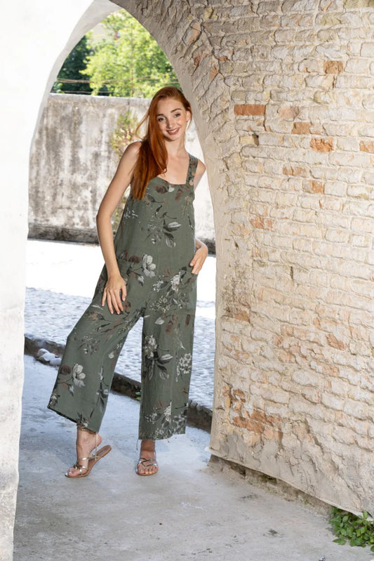 A woman with long red hair, wearing the Variations Wide-Leg Jumpsuit and sandals, stands smiling under a stone archway outdoors.