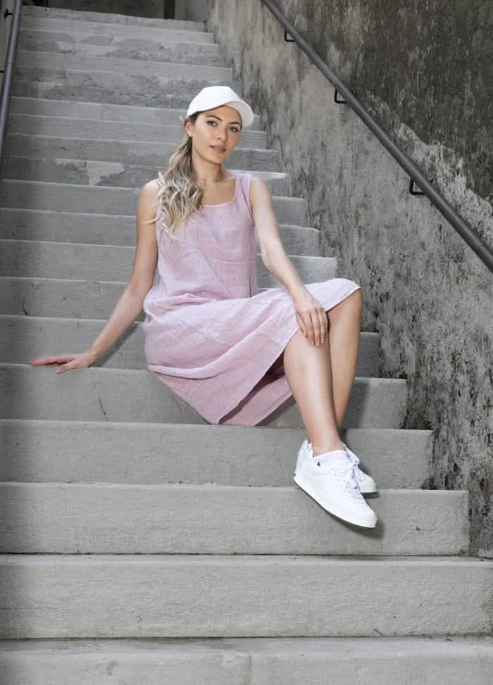 A woman wearing the Variations Linen Sleeveless Midi Dress, white sneakers, and a white cap sits on concrete stairs by a textured wall, looking at the camera.