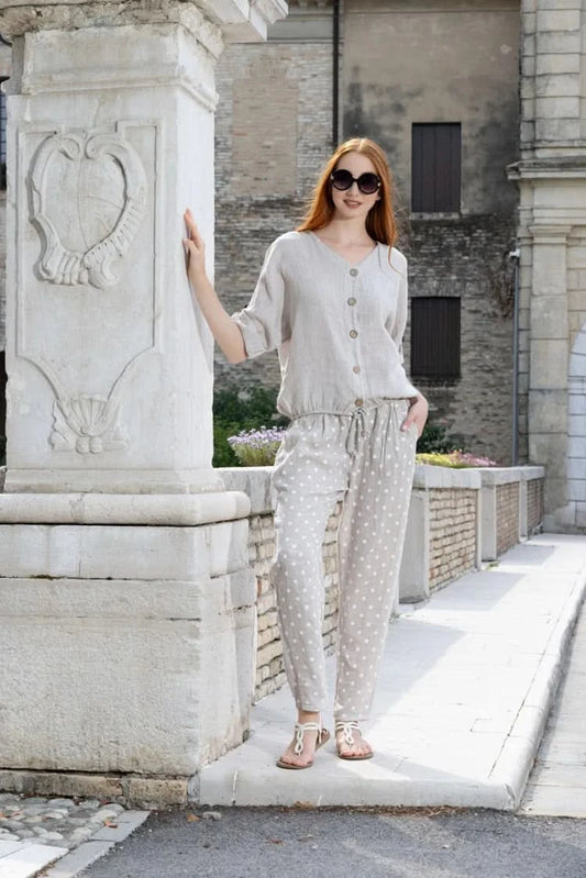 A woman stands by a stone pillar in the sun, wearing sunglasses, a light button-up shirt, and Variations Polka Dot Slim Pant.