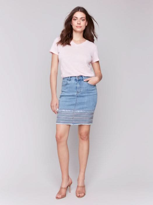 A person wears a light pink top and the Charlie B Embroidered Hem Denim Skort, paired with nude heeled sandals, standing against a plain background.