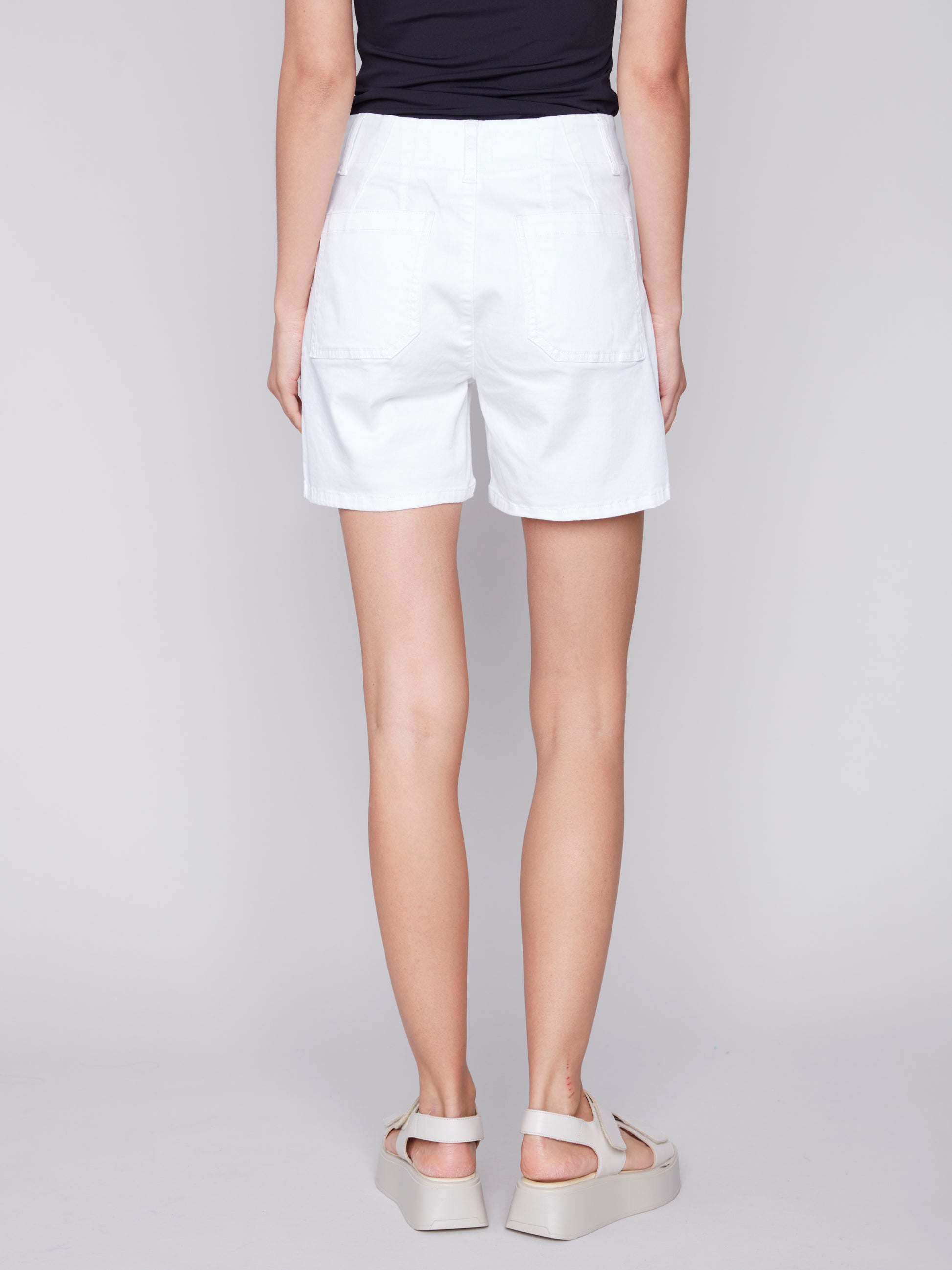 Woman showcasing a summer wardrobe in a black sleeveless top paired with Charlie B's high-quality materials white shorts with patch pocket and white sandals against a neutral background.