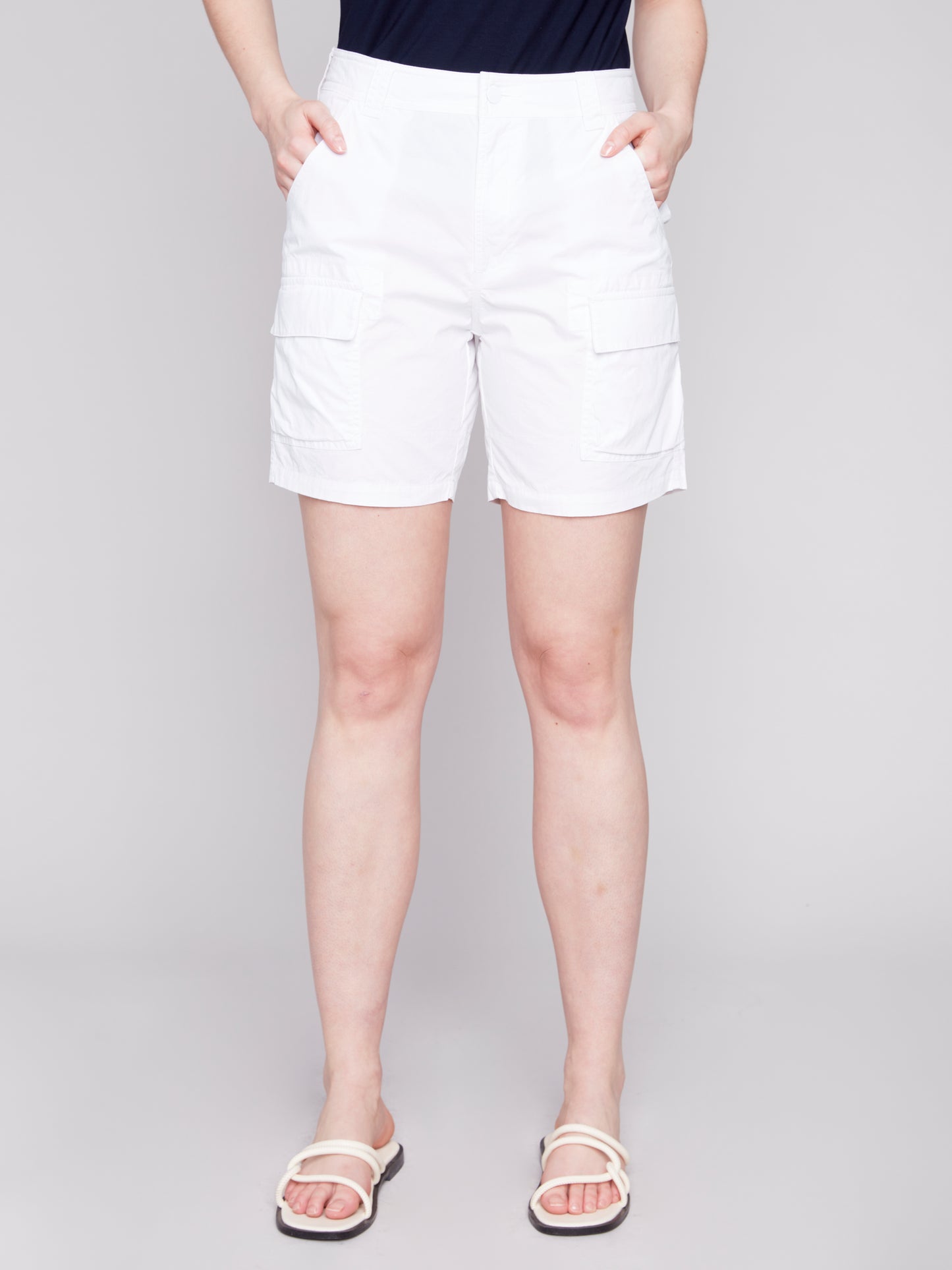 A woman stands wearing a navy tank top and stylish Charlie B Cargo Pocket Cotton Shorts with white sandals against a light gray background.