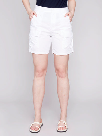 A woman stands wearing a navy tank top and stylish Charlie B Cargo Pocket Cotton Shorts with white sandals against a light gray background.