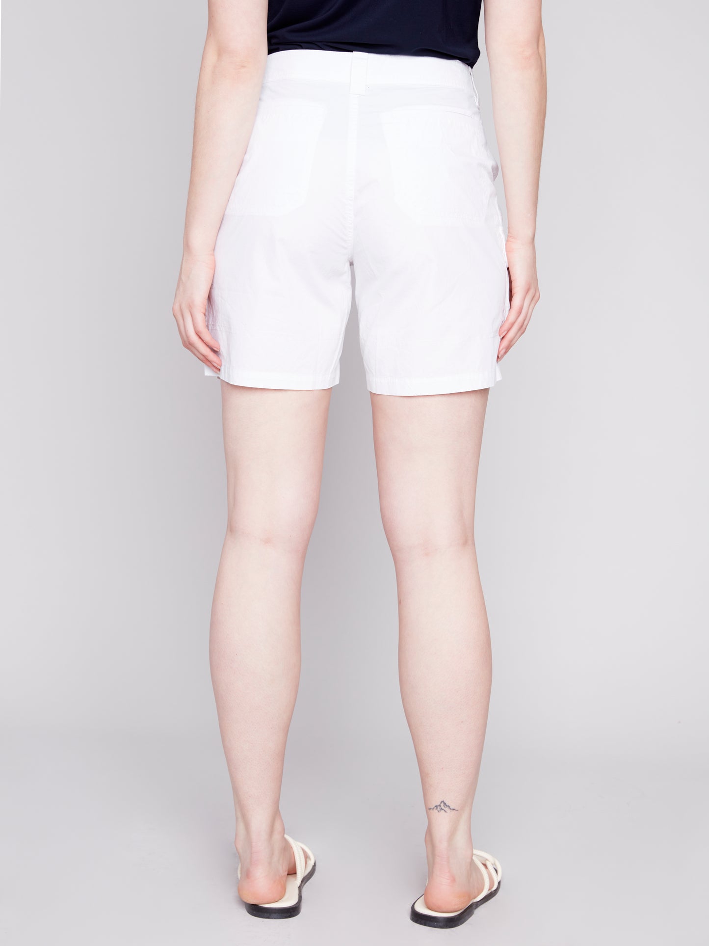 A woman stands wearing a navy tank top and stylish Charlie B Cargo Pocket Cotton Shorts with white sandals against a light gray background.