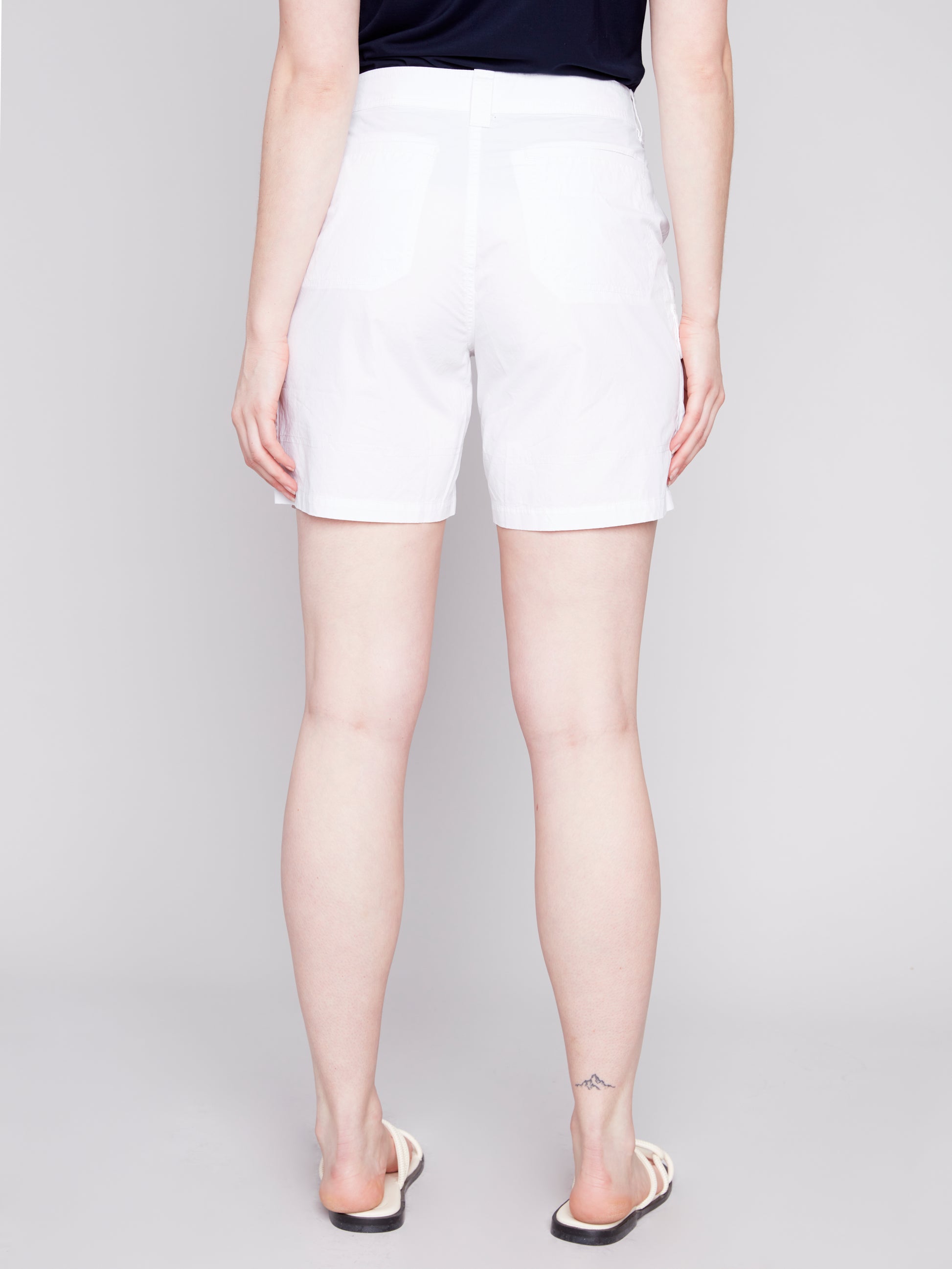 A woman stands wearing a navy tank top and stylish Charlie B Cargo Pocket Cotton Shorts with white sandals against a light gray background.