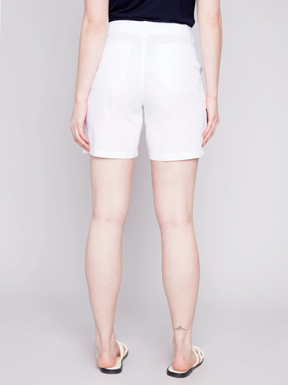 A woman stands wearing a navy tank top and stylish Charlie B Cargo Pocket Cotton Shorts with white sandals against a light gray background.