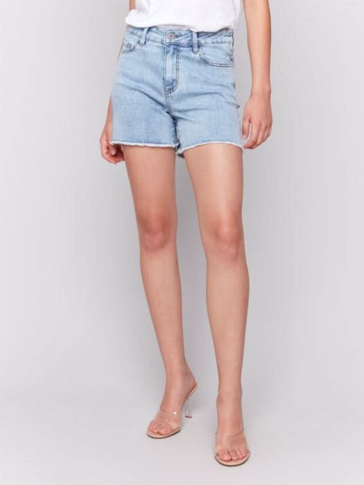 A person wears Charlie B Embellished Stretch Denim Shorts—light blue, embellished, with a frayed hem—paired with a white top and clear high-heeled sandals, standing against a plain light background.