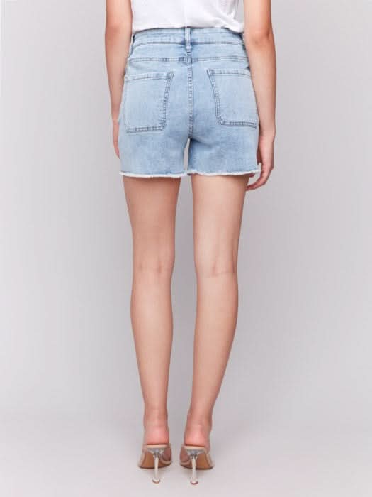 A person wears Charlie B Embellished Stretch Denim Shorts—light blue, embellished, with a frayed hem—paired with a white top and clear high-heeled sandals, standing against a plain light background.