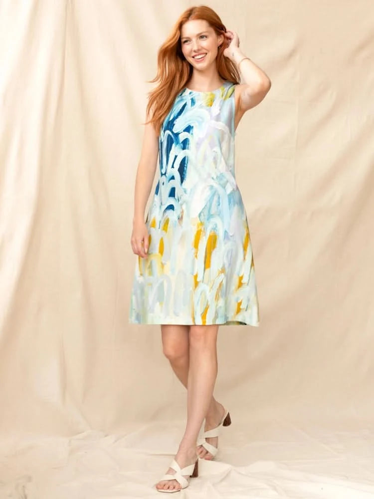 A woman with long red hair wears the Claire Desjardins Spun through the Seasons Dress—a bra-friendly, sleeveless, knee-length modal dress featuring a colorful abstract pattern—paired with white sandals against a beige fabric backdrop.