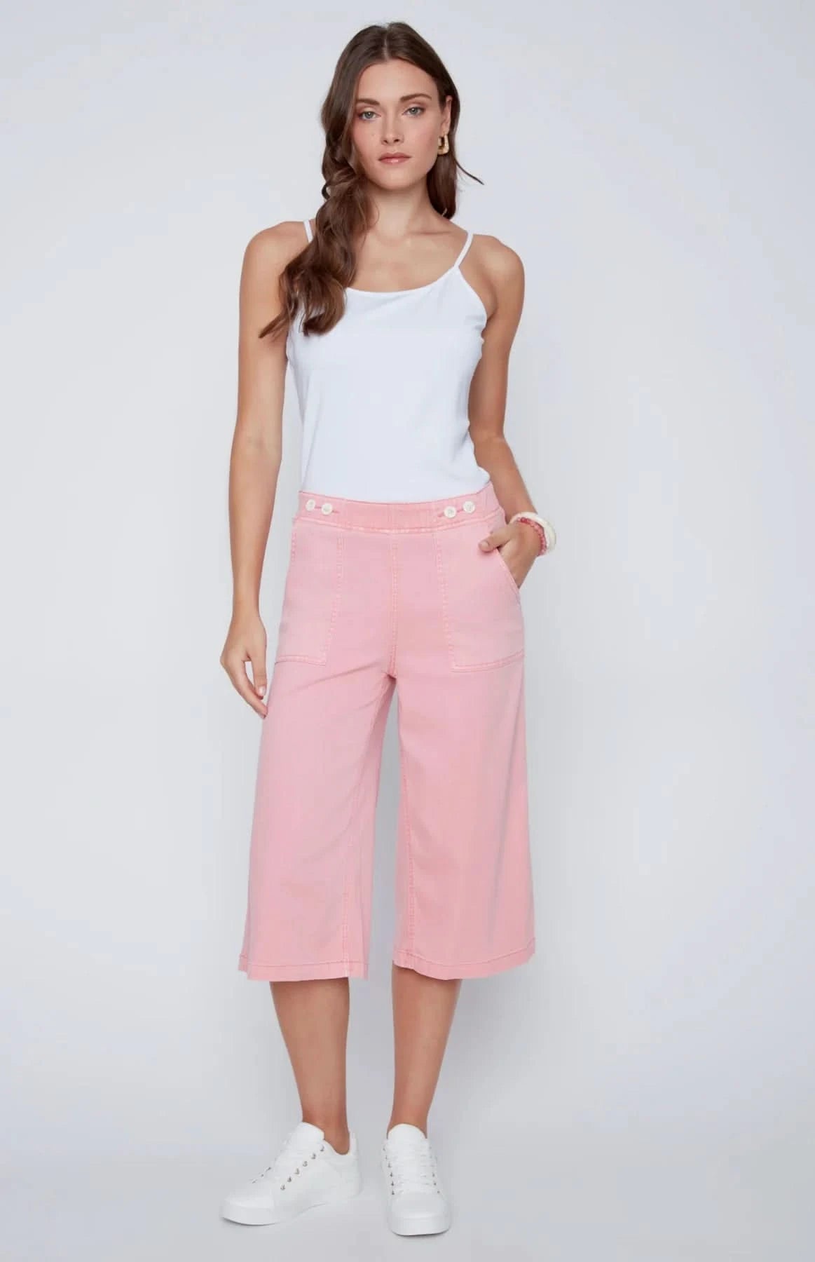 A woman stands against a plain background wearing a white sleeveless top, CoCo Y Club wide-leg cropped pants in light pink Capri Gaucho style, and white sneakers—a perfect addition to any casual wardrobe.