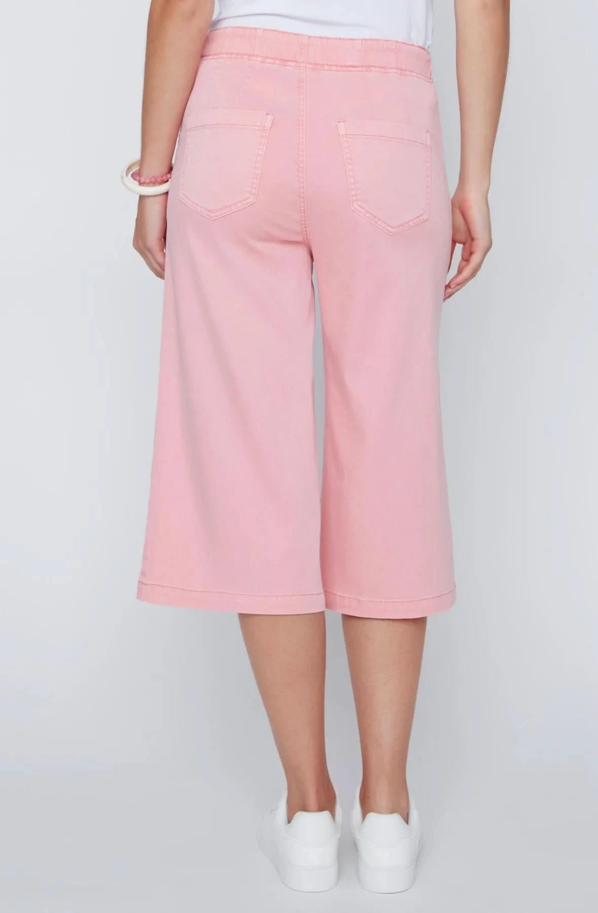 A woman stands against a plain background wearing a white sleeveless top, CoCo Y Club wide-leg cropped pants in light pink Capri Gaucho style, and white sneakers—a perfect addition to any casual wardrobe.