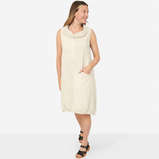 A woman stands against a white background wearing the Shannon Passero Ohio Dress, a sleeveless, light beige knee-length dress with a draped neckline, paired with black sandals.