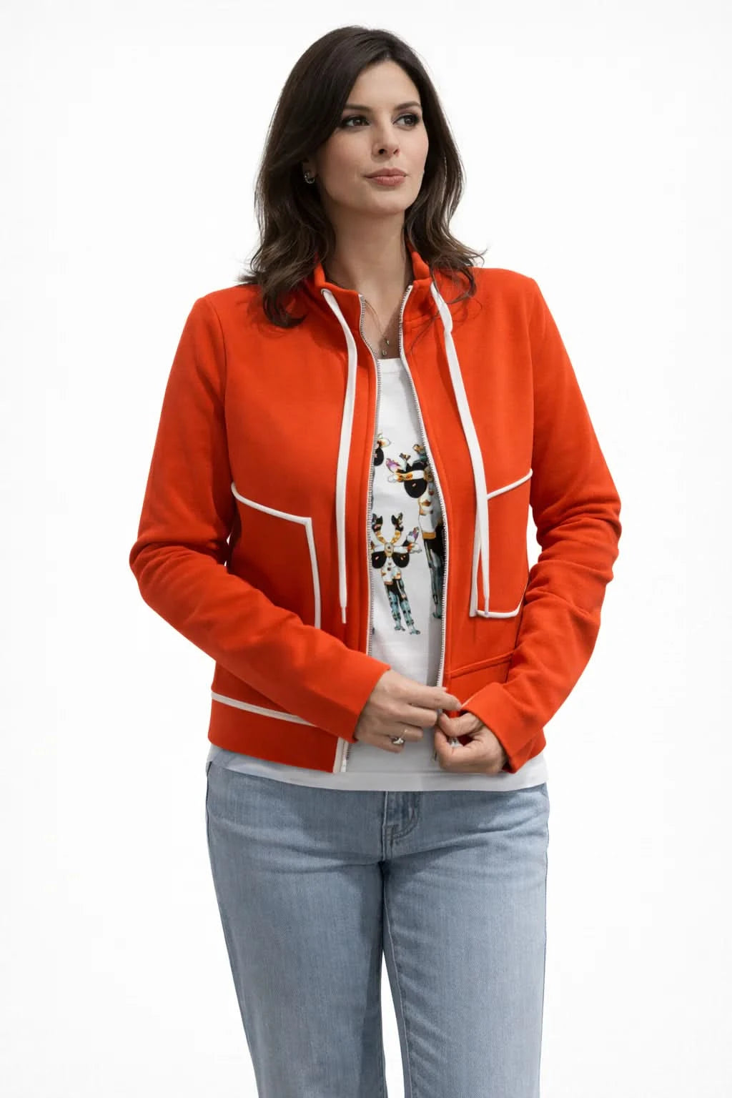 A woman wearing the Orly SPORT ZIP UP, a bright orange premium cotton cardigan with white trim, and cream pants sits on stone steps against a sunlit textured wall.