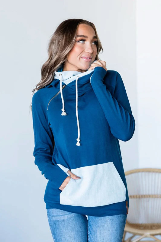 A person wearing an Ampersand Avenue Double Hooded Sweatshirt in a blue cotton blend, featuring a white pocket and drawstrings, stands indoors looking to the side, showcasing a subtle two-tone skyline design.