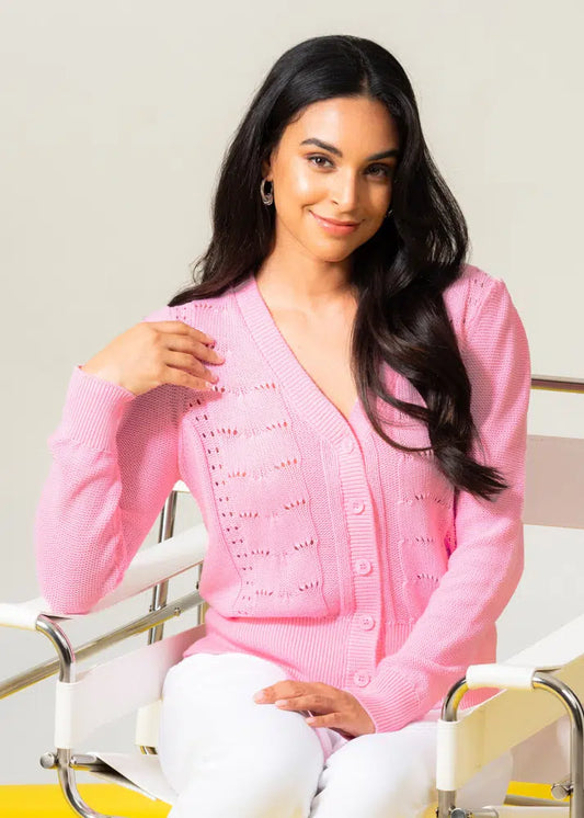 A person seated on a white chair is smiling gently while wearing Elena Wang's stylish and comfortable Front Button Knit Cardigan with a v-neckline and white pants.