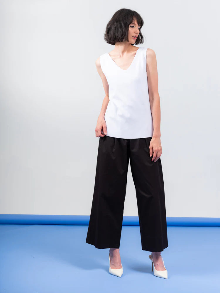 The person wears an Elena Wang Reversible Heavy Tank Top and black wide-leg pants with white heels. Posing against a blue-gray background, the fashionable top adds versatility as they look to the side.