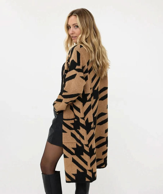 A person with long, blonde hair stands outdoors by a wooden fence, wearing a cute and soft Esqualo Jacquard Cardigan over a black shirt, leather shorts, and knee-length boots.