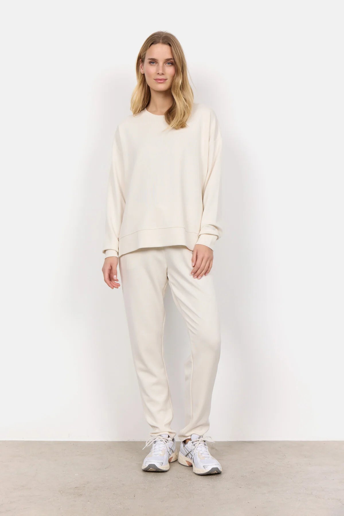 Wearing the Soya Concepts Banu Long Sleeve Sweater in off-white, paired with matching pants, this individual showcases relaxed style and all-day comfort against a simple backdrop.