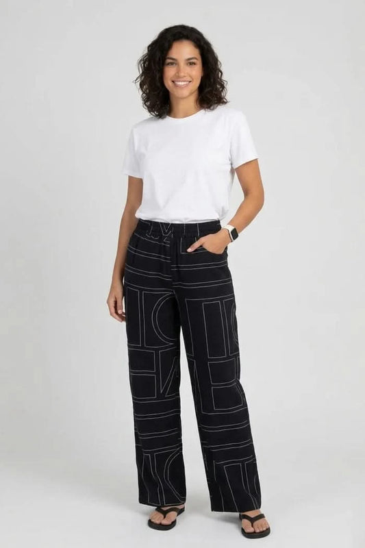 Woman standing and smiling, wearing a plain white t-shirt and Shannon Passero Lani Pant in breathable printed rayon, paired with black sandals and a smartwatch, against a plain light background.