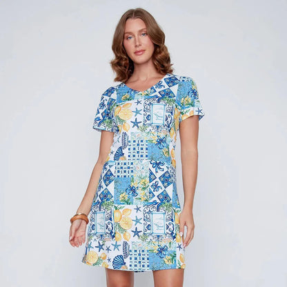 A woman stands smiling in the CoCo Y Club ‘Amalfi’ Lace Detail Dress, a short sleeve style featuring blue and yellow Greek-inspired tile and lemon motifs on a white background.