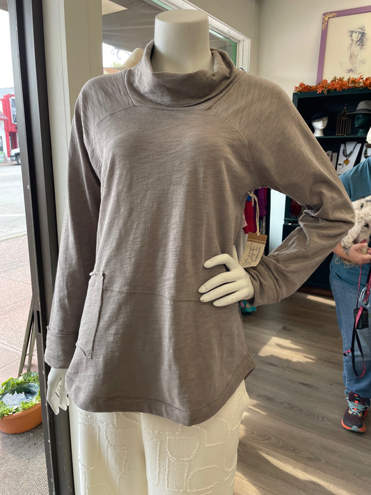 A mannequin in a retail store showcases the Escape Weekend cowl, a long-sleeve light brown cowl neck top with front pockets for cozy comfort, paired with a white textured skirt.