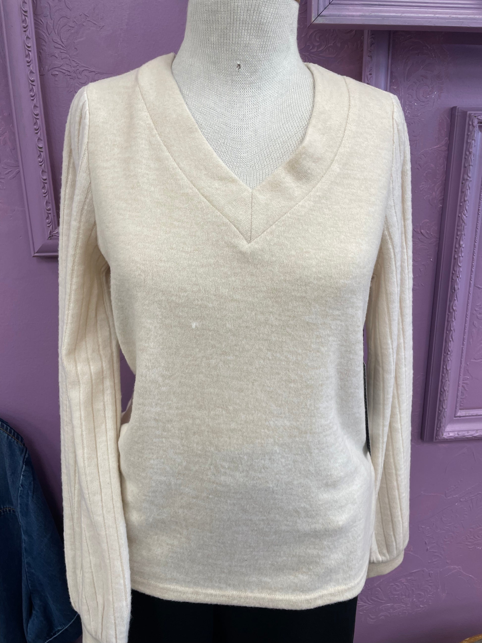 The Cream sweater Point Zero by Motion, featuring long sleeves and a V-neck, is shown on a mannequin in front of a lavender wall with decorative molding.
