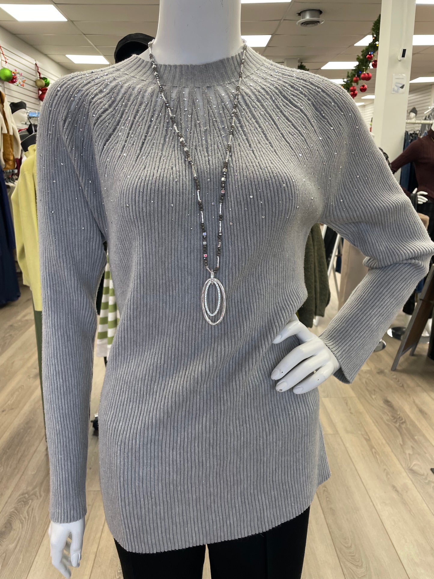A mannequin in a retail store showcases the Nu-Look Fashions High Neck L/S Top, a light gray ribbed long-sleeve sweater with gem details, accessorized with a long silver necklace.