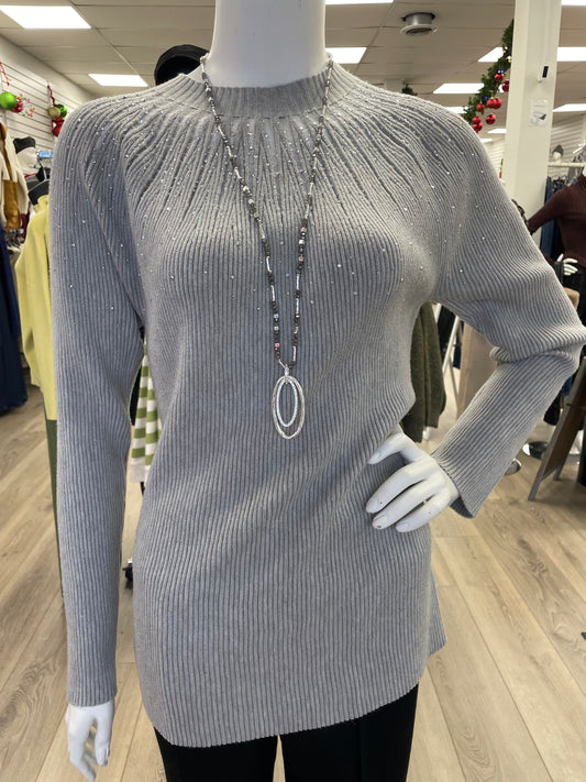 A mannequin in a retail store showcases the Nu-Look Fashions High Neck L/S Top, a light gray ribbed long-sleeve sweater with gem details, accessorized with a long silver necklace.