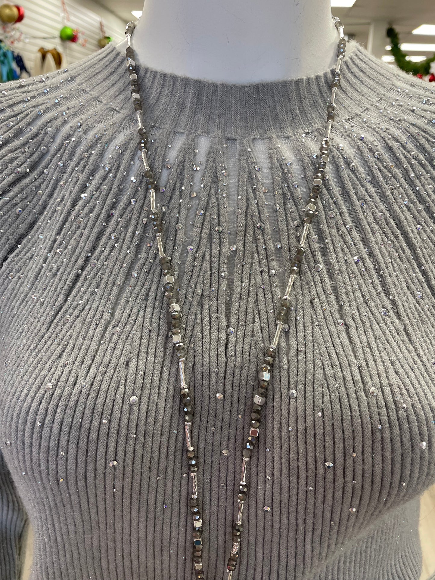 A mannequin in a retail store showcases the Nu-Look Fashions High Neck L/S Top, a light gray ribbed long-sleeve sweater with gem details, accessorized with a long silver necklace.